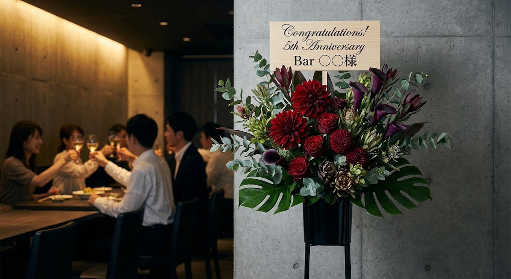 コンクリート打ちっぱなしのおしゃれなバーに届いた、深みのある赤とグリーンを基調としたシックな周年祝いのスタンド花。背景には乾杯する客席の様子。