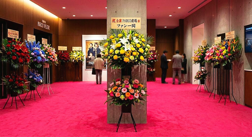 渋谷パルコ劇場のロビーに並ぶ、色とりどりの公演祝いスタンド花。特徴的なショッキングピンクの絨毯に、黄色、赤、青、ピンクなど多彩な花々が美しく映え、舞台初日の華やかな活気を演出している様子。
